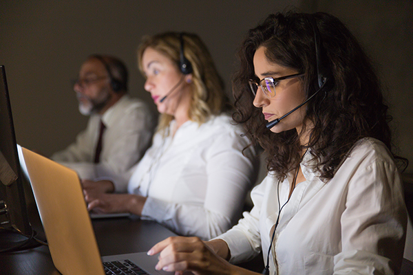 Team of teleworkers working in dark office. Side view of professional business people in headsets working with computers in dark office. Working late concept